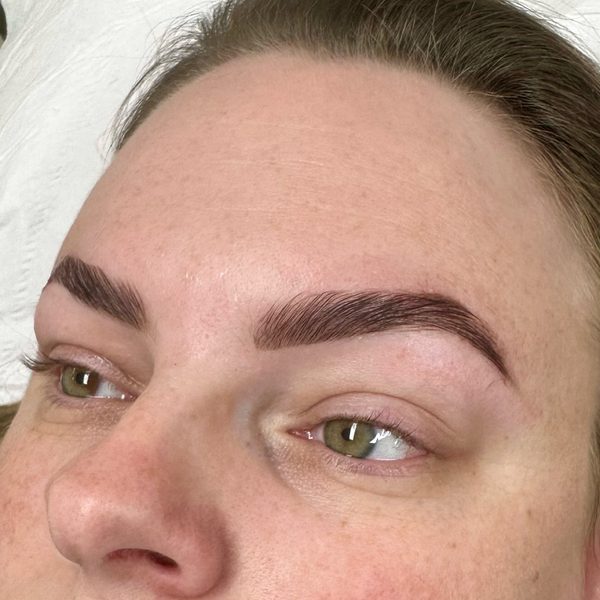 Close-up of brow lamination with stain and wax on fair, freckled skin: reddish-brown brows brushed upward and shaped to a clean edge.