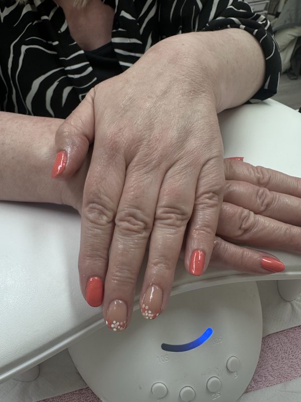 Short almond builder gel nails in coral resting on a white nail lamp, with one nude accent nail hand-painted with small white daisies.